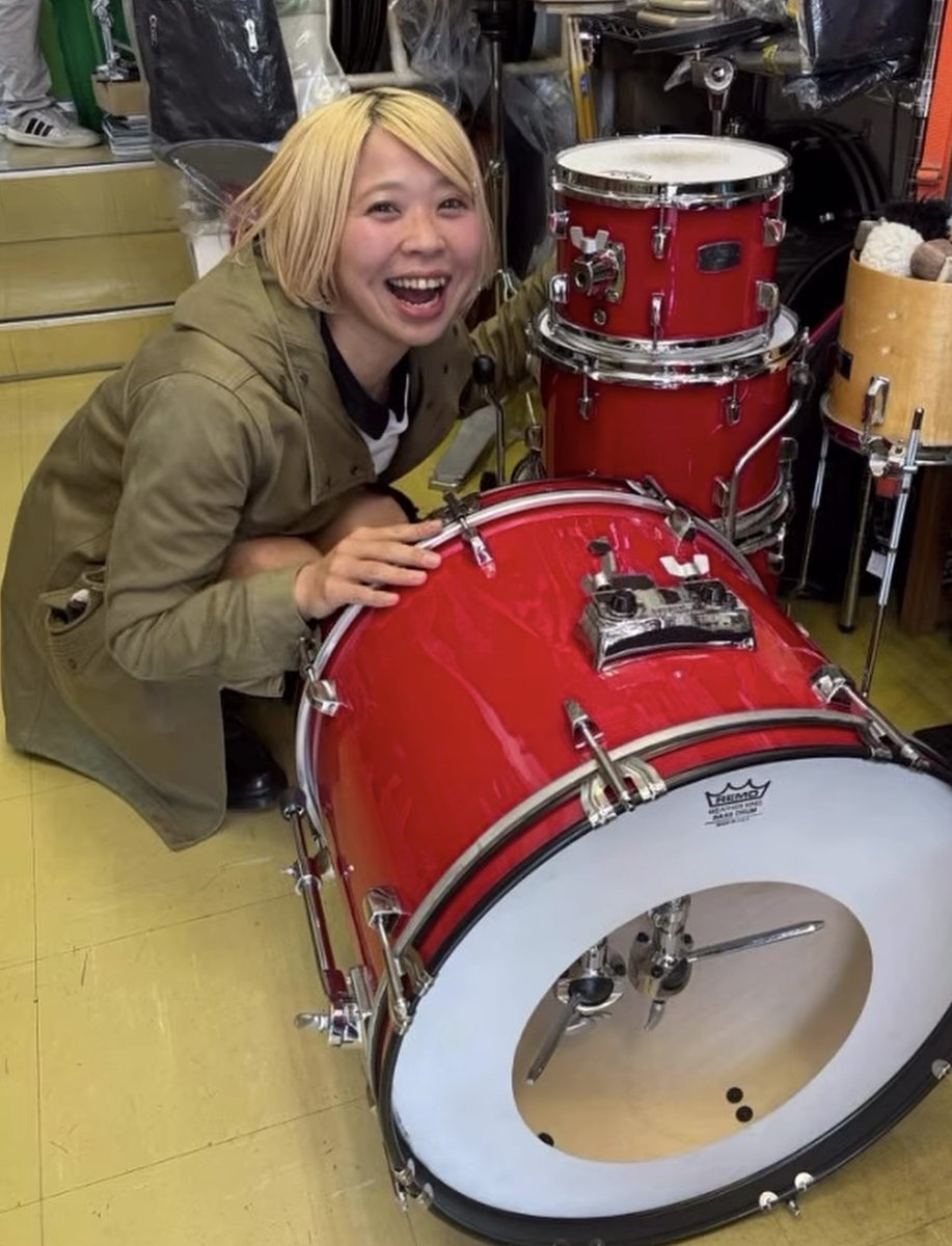 赤いドラムセットの横で、笑顔でしゃがむ人